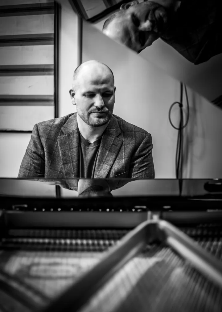 Maxwell Henry seated at a grand piano, photographed from inside the instrument, his reflection visible in the raised lid